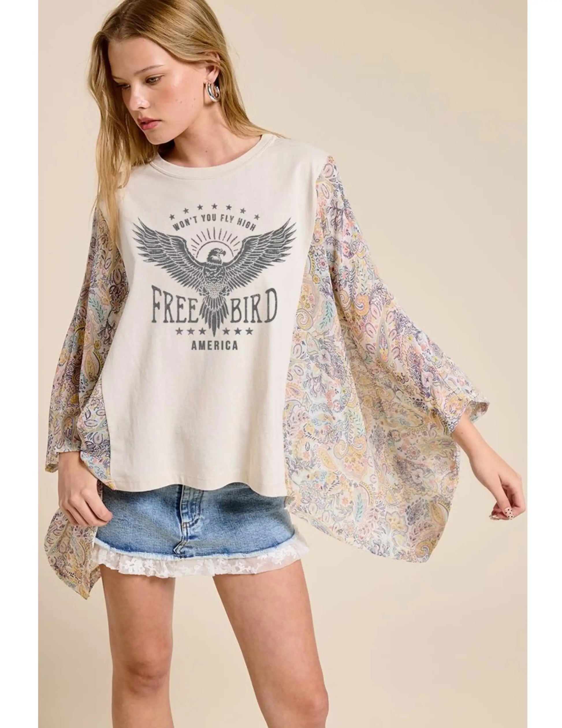 Woman wearing a graphic t-shirt with an eagle design and  floral kimono sleeves and denim shorts on a beige background.