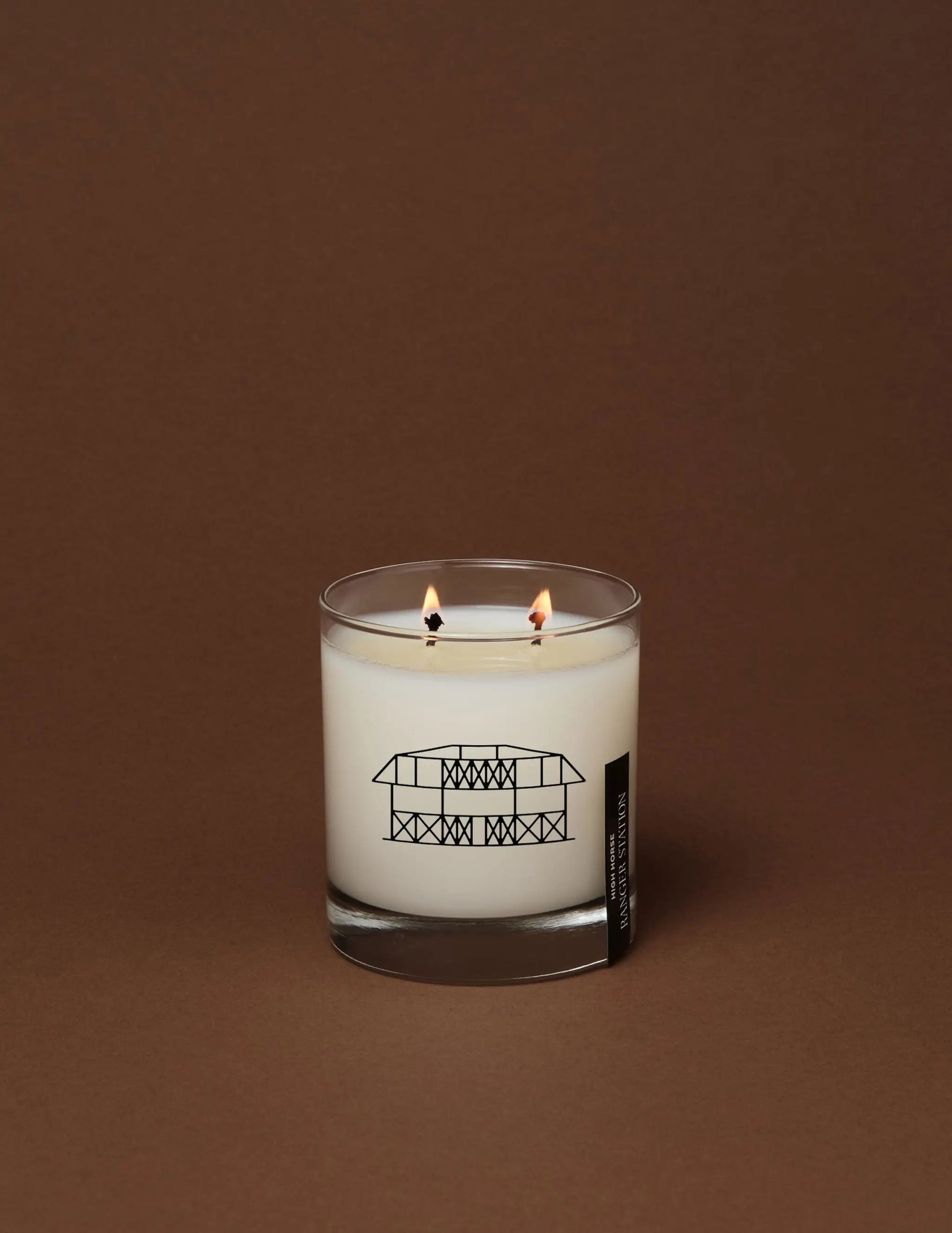 A white scented candle in a clear glass jar with two lit wicks and a black building line drawing on the opry 100 ranger station high horse candle